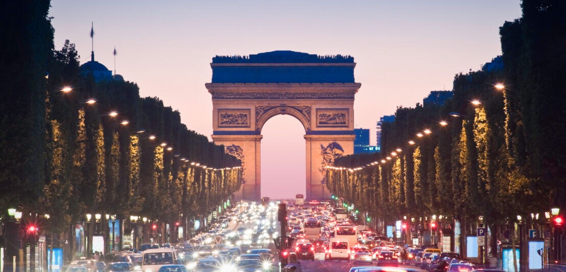 Reger Autoverkehr vor dem beleuchteten Arc de Triomphe in Paris am Abend.