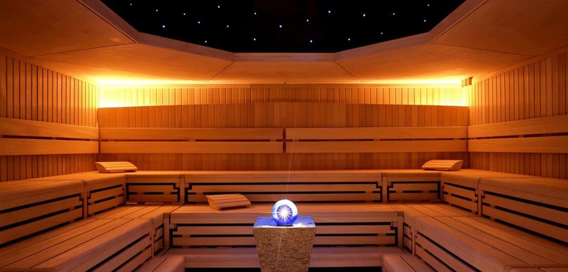 Das Innere einer Holzsauna mit Glaskugel auf einem Podest im Zentrum und einem angedeuteten, beleuchteten Sternenhimmel an der Decke