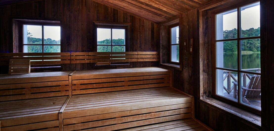 Das Innere einer Holzsauna, durch deren Fenster ein See und Waldstück zu erkennen ist