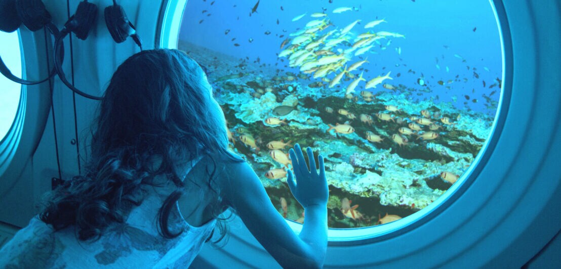Ein Mädchen schaut durch ein Bullauge in einem U-Boot auf Fische und Korallen.