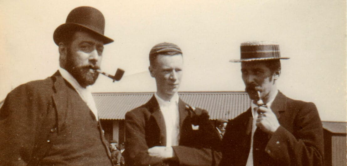Ein historisches Foto dreier Männer, zwei tragen Hüte und rauchen Pfeife