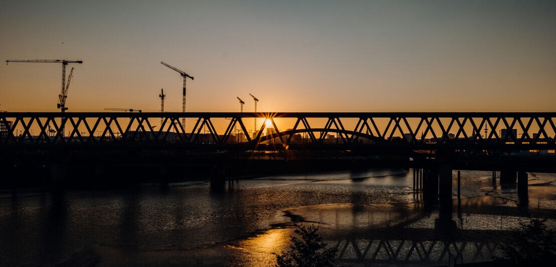 Blick auf einen Fluss, eine Eisenbrücke und Kräne bei Sonnenuntergang