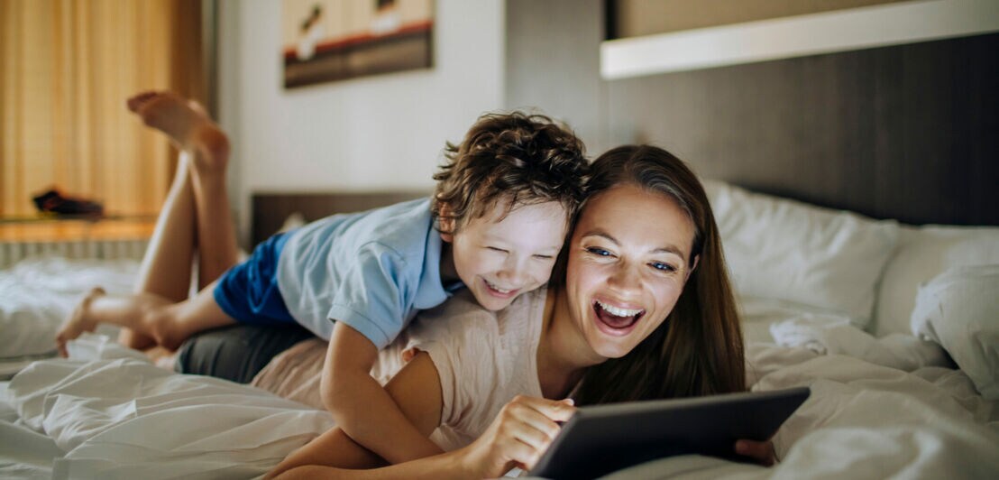 Eine Mutter und ihr kleiner Sohn liegen aneinander gekuschelt im Bett und schauen lachend auf ein Tablet