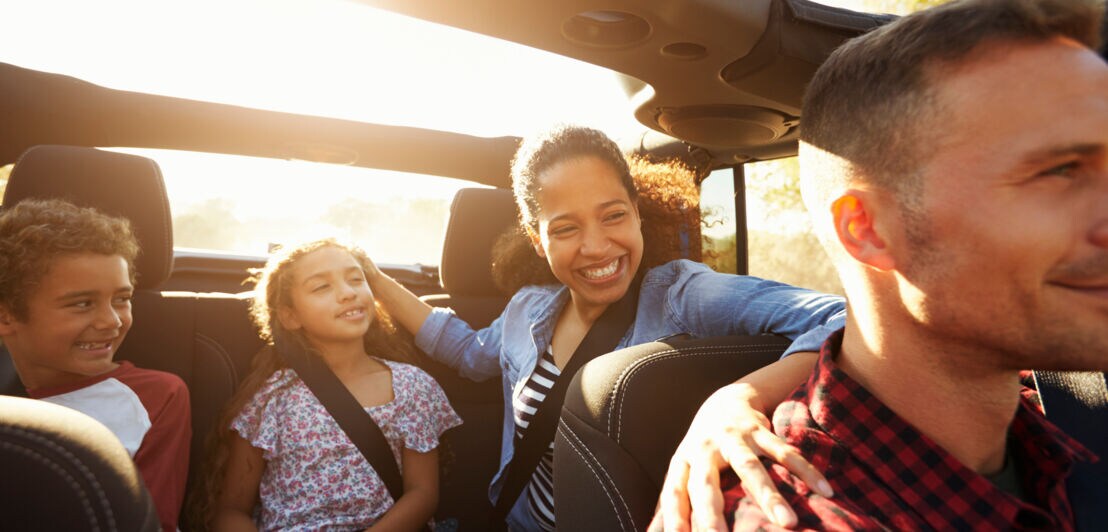 Eine lachende Frau und zwei Kinder auf dem Rücksitz eines Cabriolets blicken nach vorne zum Fahrer des Autos 
