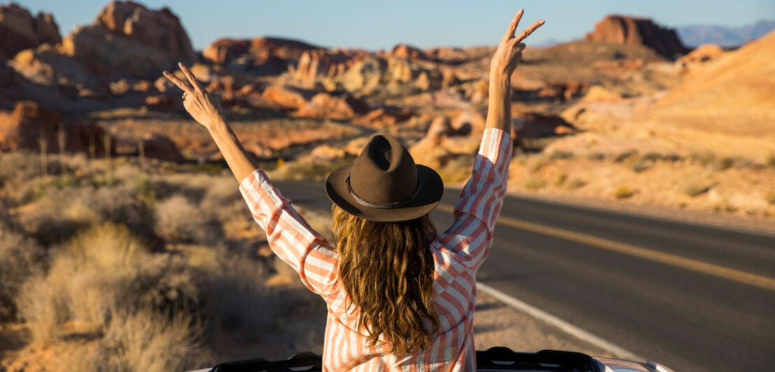 Rückansicht einer Frau mit Hut, die auf einer Reise durch eine Wüstenlandschaft mit dem Oberkörper aus dem Schiebedach eines Mietwagens schaut und die Hände in die Luft streckt