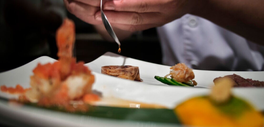 Die Hand eines Küchenchef beträufelt mit einem Löffel ein exquisites Gericht auf einem weißen Teller