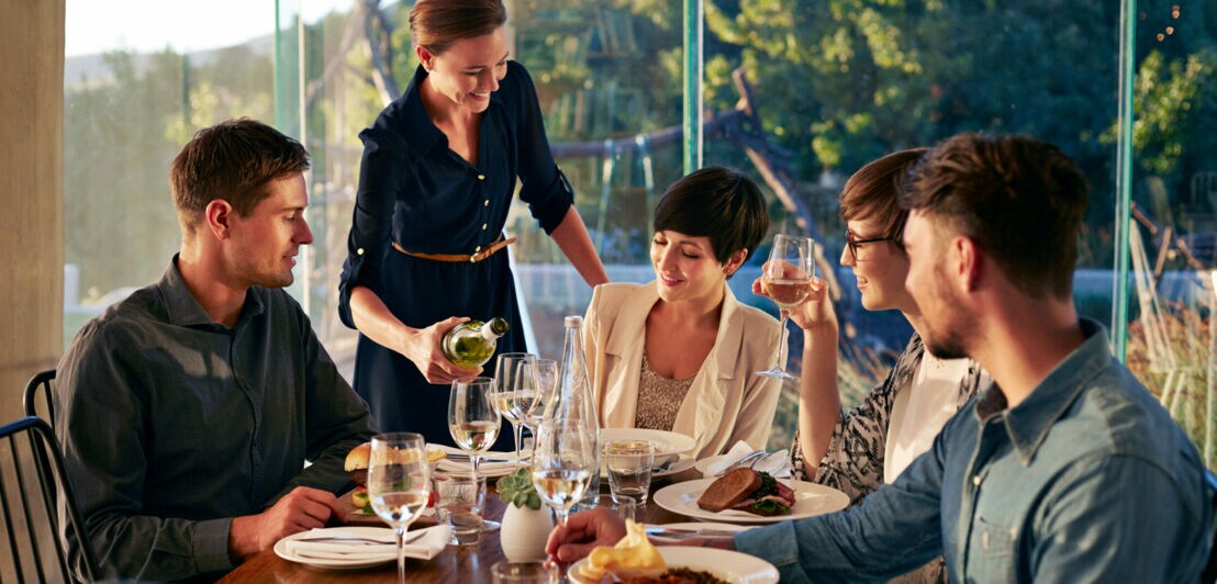 Eine Kellnerin gießt Wein in ein Glas an einem elegant gedeckten Tisch mit einer Gruppe junger Menschen