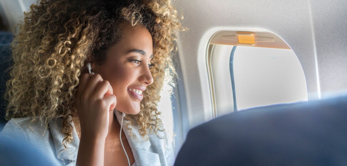 Nahaufnahme einer jungen, lächelnden Frau mit Kopfhörern auf einem Fensterplatz in einem Flugzeug während der Reise