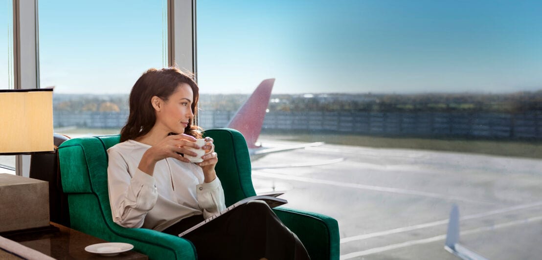 Eine elegante Frau sitzt mit einer Tasse Kaffee in einer Flughafen-Lounge und blickt durch ein Panoramafenster aufs Rollfeld