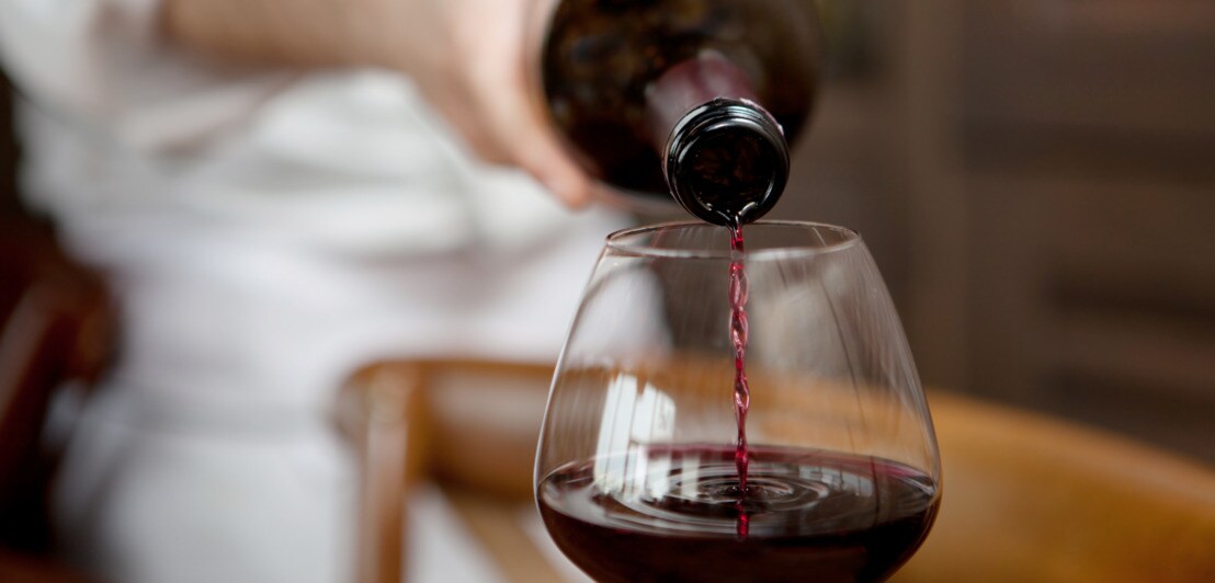 Eine Person im weißen Hemd schenkt Rotwein in ein bauchiges Weinglas