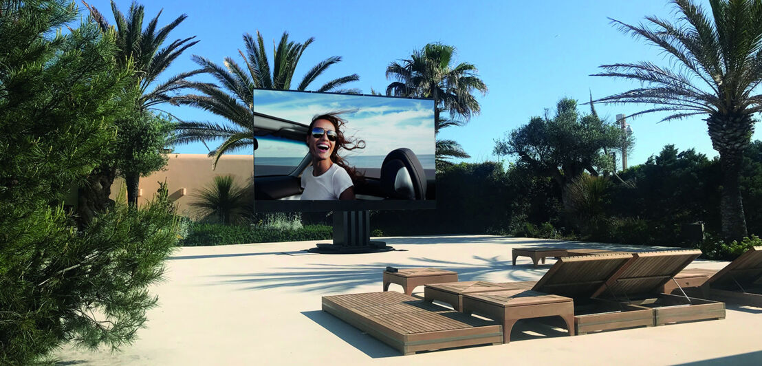 Der Fernseher C SEED 201 in einem großzügigen Terrassenbereich und von Palmen umgeben.