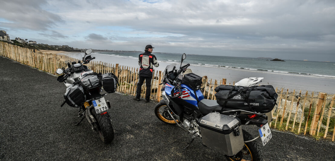 Zwei Motorräder parken vor einem Zaun, im Hintergrund zeichnen sich das Meer und die Umrisse von Saint-Malo ab.