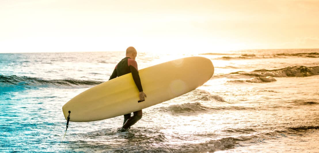 Ein Surfer trägt sein Surfboard durch das Wasser im Sonnenuntergangslicht.