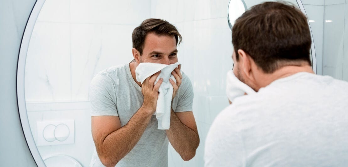 Ein Mann trocknet sich im Badezimmer sein Gesicht mit einem Handtuch ab und blickt dabei in den Spiegel.