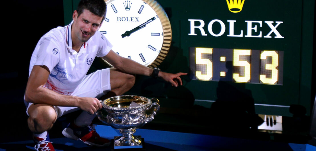 Ein Tennisspieler mit einem Pokal zeigt auf eine Uhr