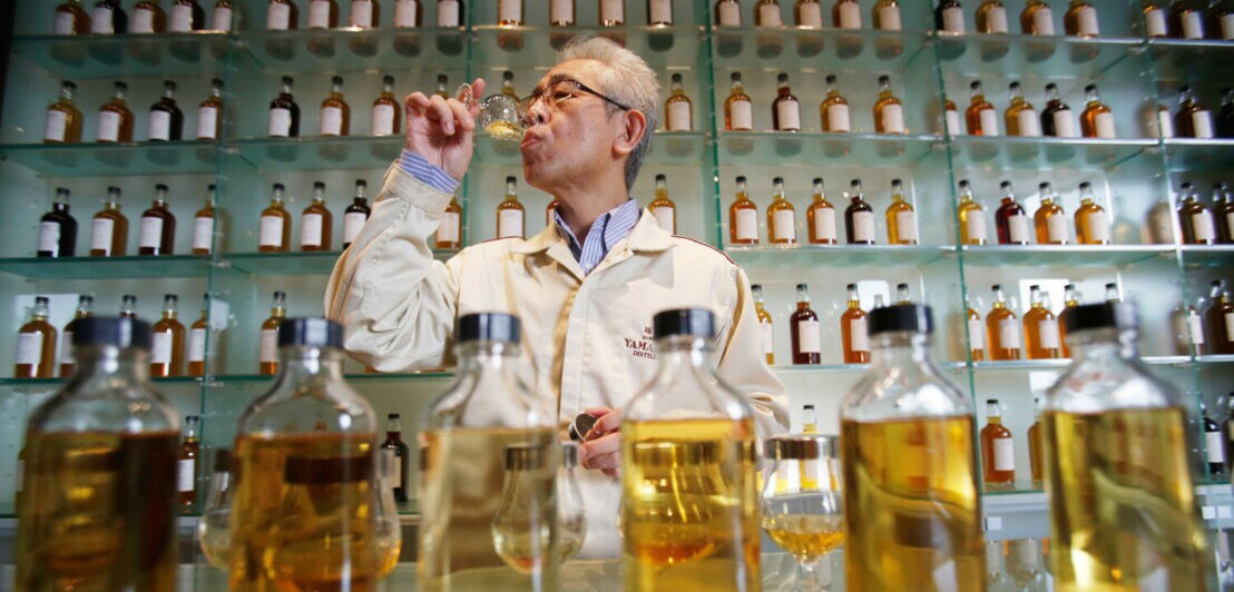 Ein Mann in einem weißen Kittel verkostet Whisky in einer Destillerie vor einem Regal mit Flaschen