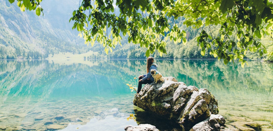 Eine Frau sitzt mit einem Hund auf einem Felsen an einem klaren Bergsee