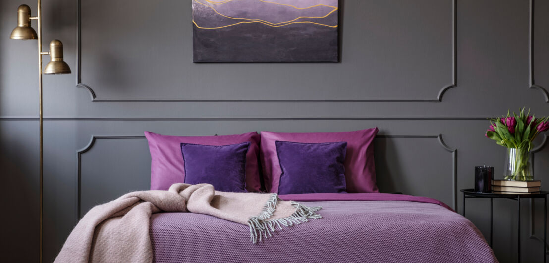 Ein elegantes Schlafzimmer mit einem Doppelbett mit Kissen und Decke in Violetttönen vor einer grauen Wand