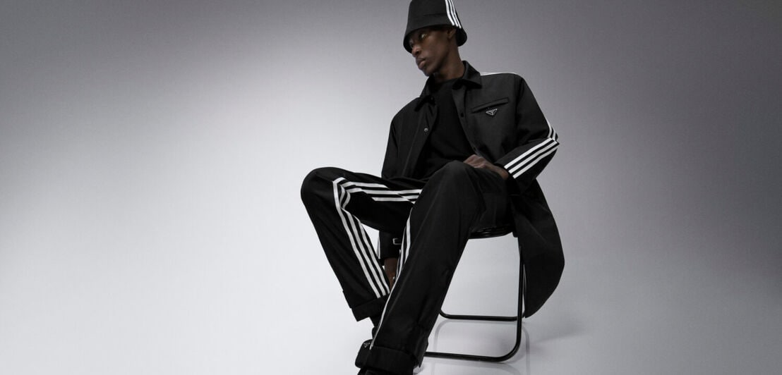 Ein männliches Model mit schwarzem Trainingsanzug von adidas sitzt auf einem Stuhl.