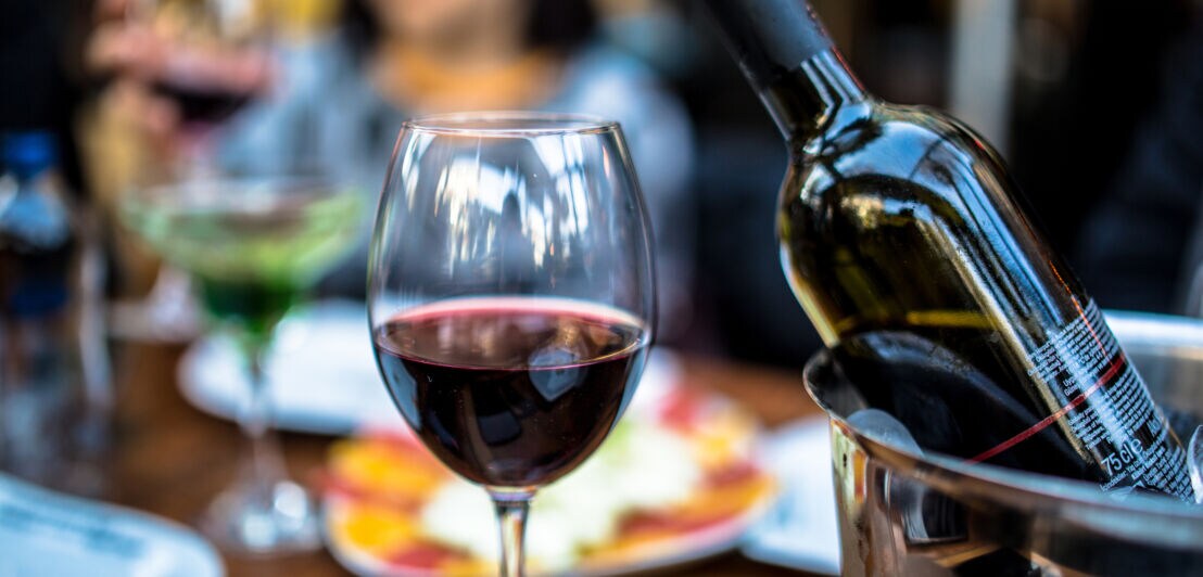 Ein gefülltes Glas Rotwein neben einem Weinkühler.