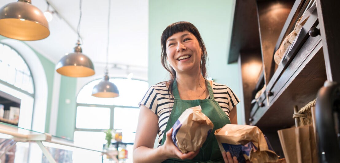 Eine Verkäuferin mit Backwaren in den Händen steht lachend hinter der Theke einer Bäckerei
