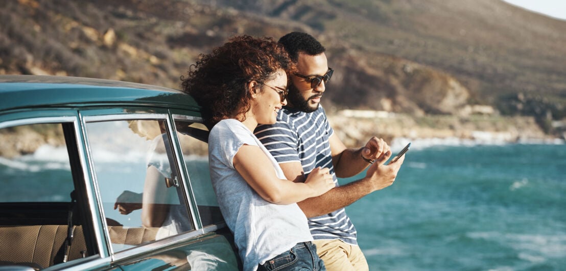 Ein Paar in Freizeitkleidung lehnt sich an ein Auto an einer Küste und schaut gemeinsam auf ein Smartphone