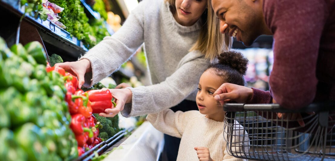 Ein kleines Mädchen steht mit seinen Eltern im Supermarkt vor dem Gemüseregal und sucht Paprika aus
