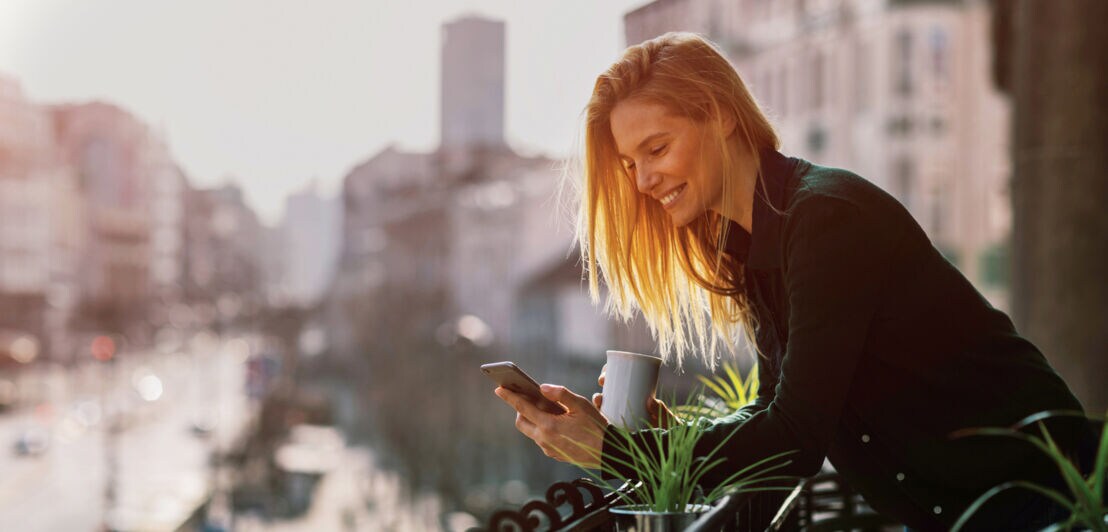 Eine Frau steht mit einer Kaffeetasse auf einem Balkon und schaut auf ihr Smartphone