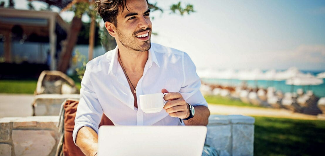 Ein junger Mann im weißen Hemd sitzt an einem Strand mit einer Kaffeetasse vor einem Laptop in der Sonne