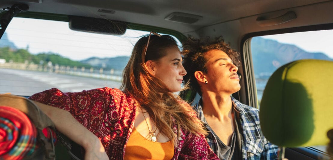 Ein junges, reisendes Paar schaut vom Rücksitz eines Autos in die Abendsonne