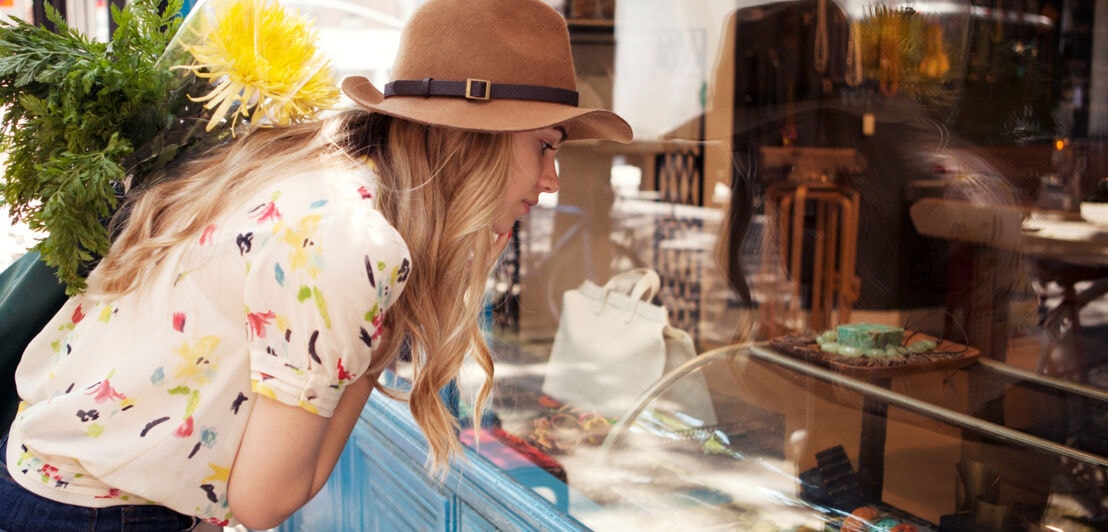 Eine junge Frau mit Hut und Einkäufen im Arm betrachtet die Auslage im Schaufenster einer Boutiqu