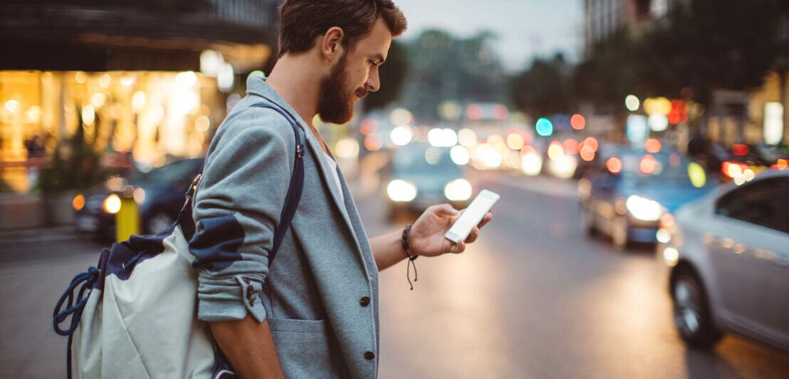 Ein junger Mann mit Rucksack steht an einer befahrenen Straße und schaut auf sein Smartphone