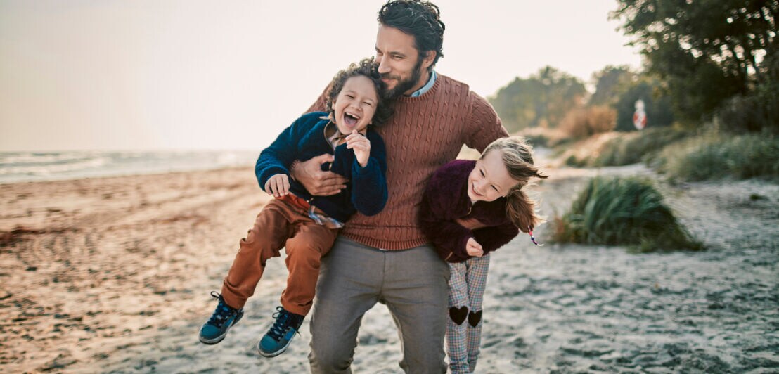 Ein Mann trägt unbeschwert zwei kleine, lachende Kinder unter dem Arm
