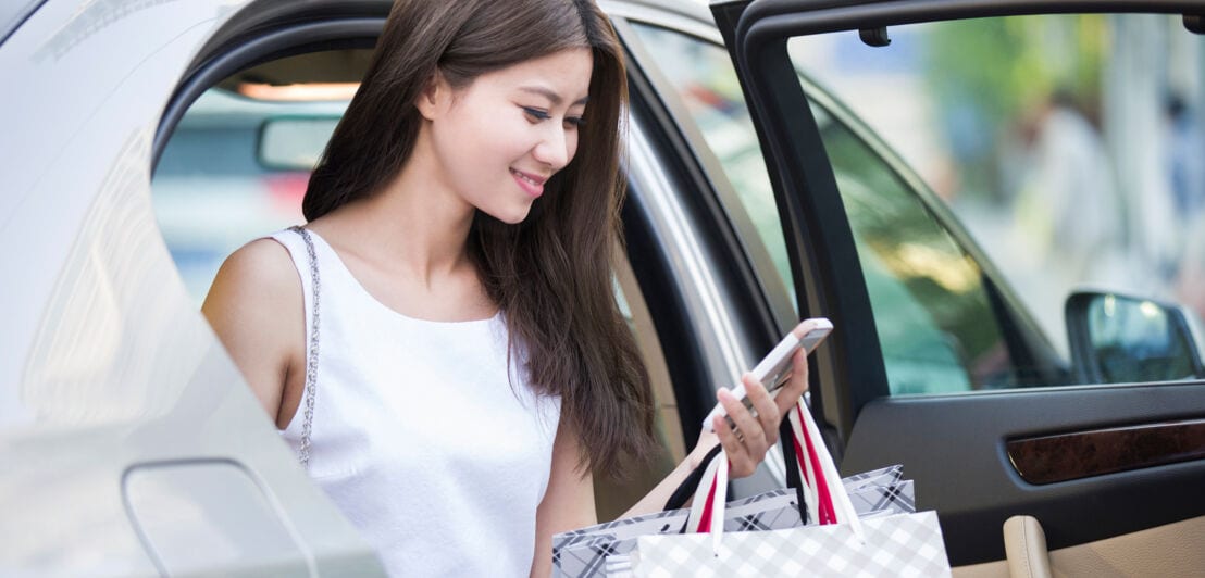 Eine elegante, junge Frau mit Einkaufstaschen in der Hand steigt aus einem Auto und schaut lächelnd auf ihr Smartphone 