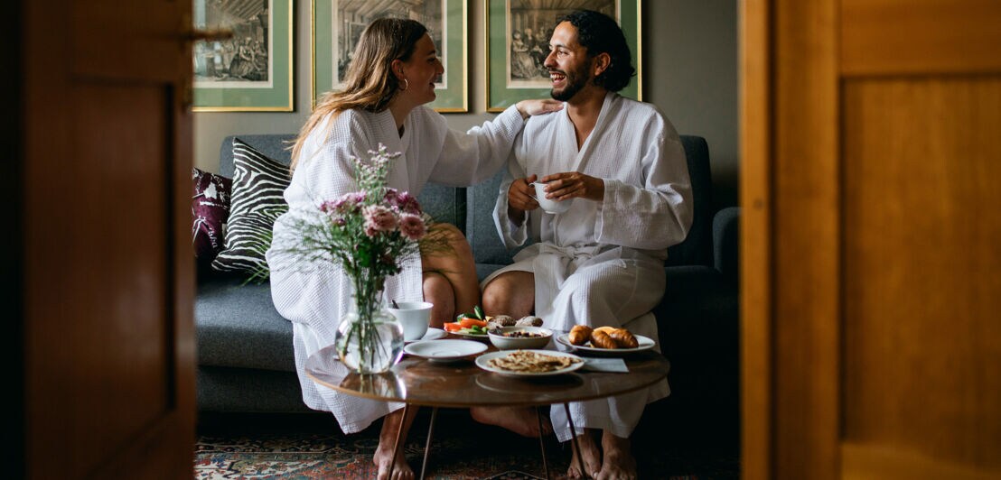 Ein Paar sitzt gut gelaunt in weißen Bademänteln auf einem Sofa in einer Hotelsuite und genießt das gemeinsame Frühstück