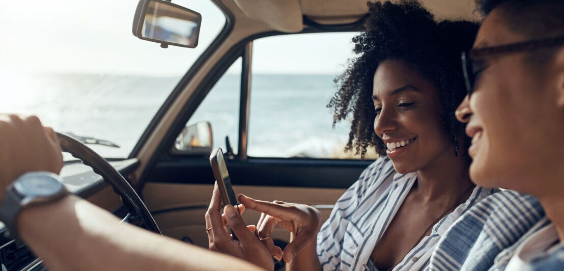 Zwei Frauen in einem Auto an der Meeresküste schauen lächelnd auf ein Smartphone