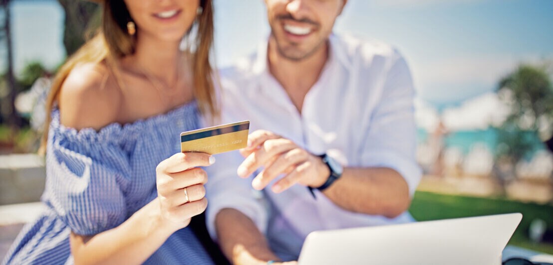 Mann und Frau sitzen im Urlaub vor einem Laptop und blicken gemeinsam auf eine Kreditkarte