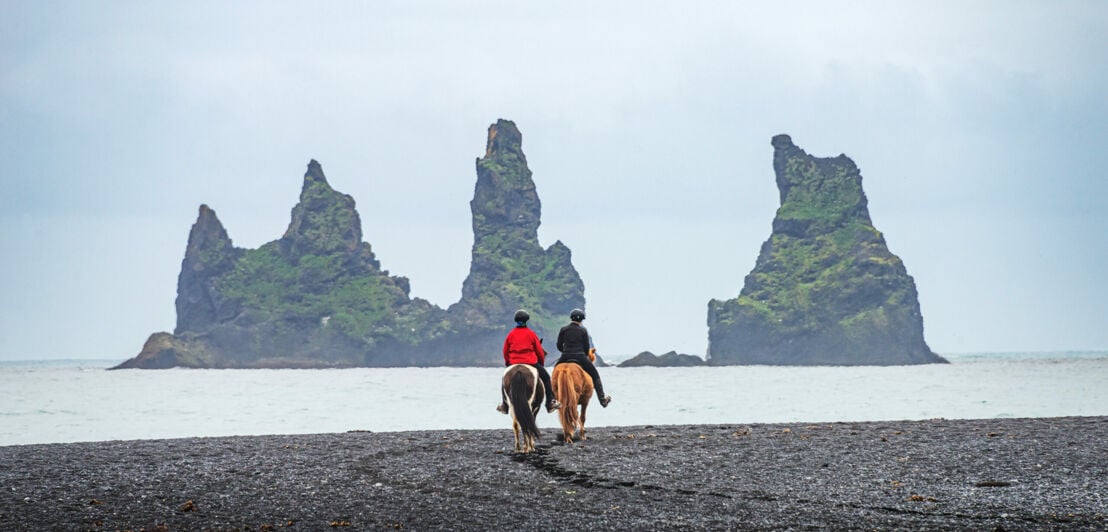 Zwei Menschen auf Pferden vor den Lavasäulen nahe Vík in Südisland