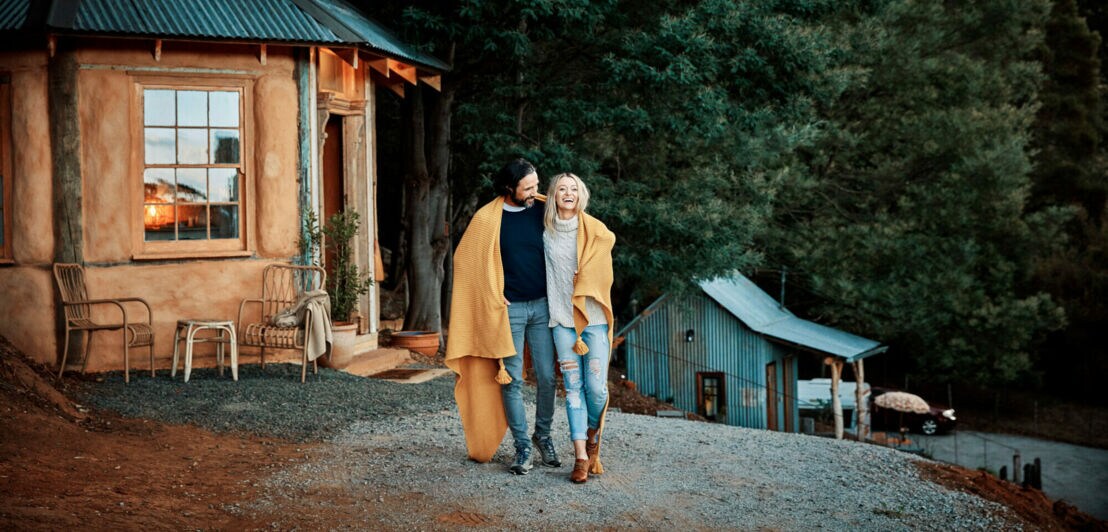 Ein glückliches, stylishes Paar steht in eine Decke gekuschelt vor einer gemütlichen Holzhütte in einem Waldgebiet