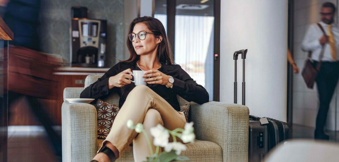 Eine Frau mit einer Kaffeetasse in der Hand sitzt entspannt in einer Airport-Lounge