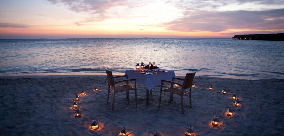 Ein gedeckter Tisch am Strand eines Luxusresorts, umgeben von Kerzen bei Sonnenuntergang