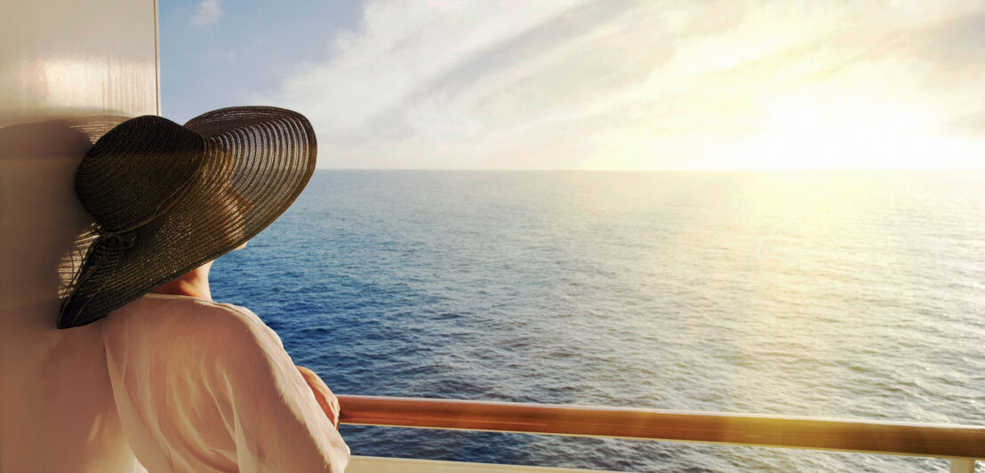 Eine elegante Frau mit Sonnenhut schaut im Sonnenuntergang von einem privaten Balkon eines Kreuzfahrtschiffes auf das offene Meer hinaus