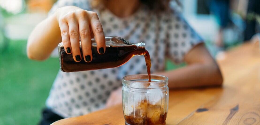 Eine junge Frau gießt kalten Kaffee in ein Glas mit Eiswürfeln ein.