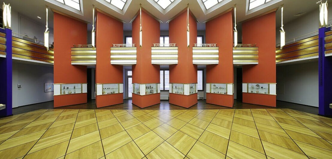 Ein geometrisch angelegter Raum mit Holzfußboden, orangen Säulen und einer Empore