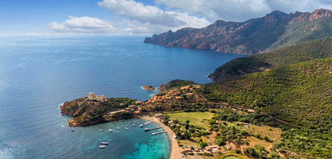 Blick über eine Bucht auf Korsika mit türkisem Wasser, ein paar Booten und einer Gebirgskette im Hintergrund.