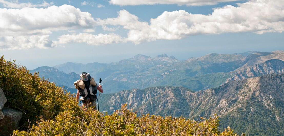Ein Mann wandert in großer Höhe über einen Berg, dazu Panoramablick über das Gebirge.