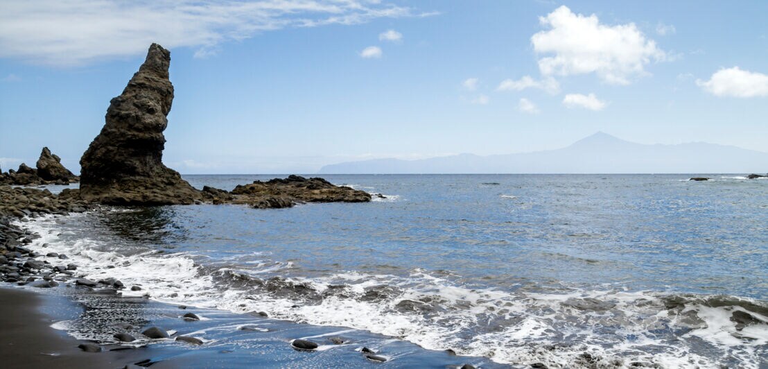 Sandstrand mit Steinen und Felsformationen, am Horizont ist der Teide auf Teneriffa zu erkennen.