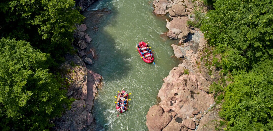 Luftaufnahme zweier Raftingboote in einem Fluss zwischen Felsen und Bäumen