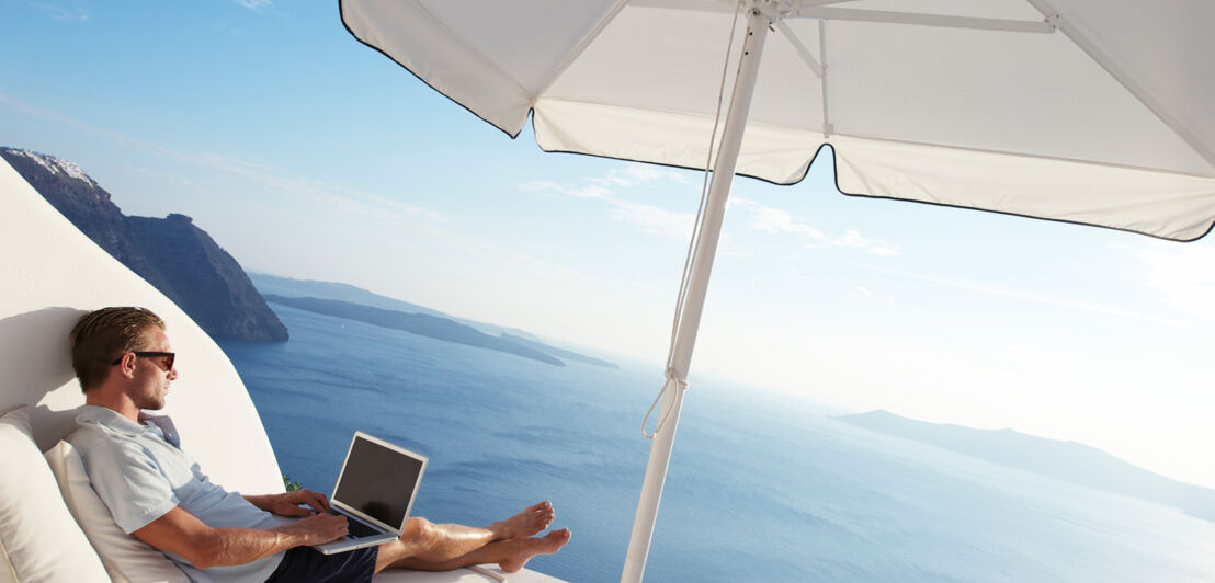 Ein leger gekleideter Mann mit Laptop auf dem Schoß auf einer luxuriösen Terrasse mit Meerblick
