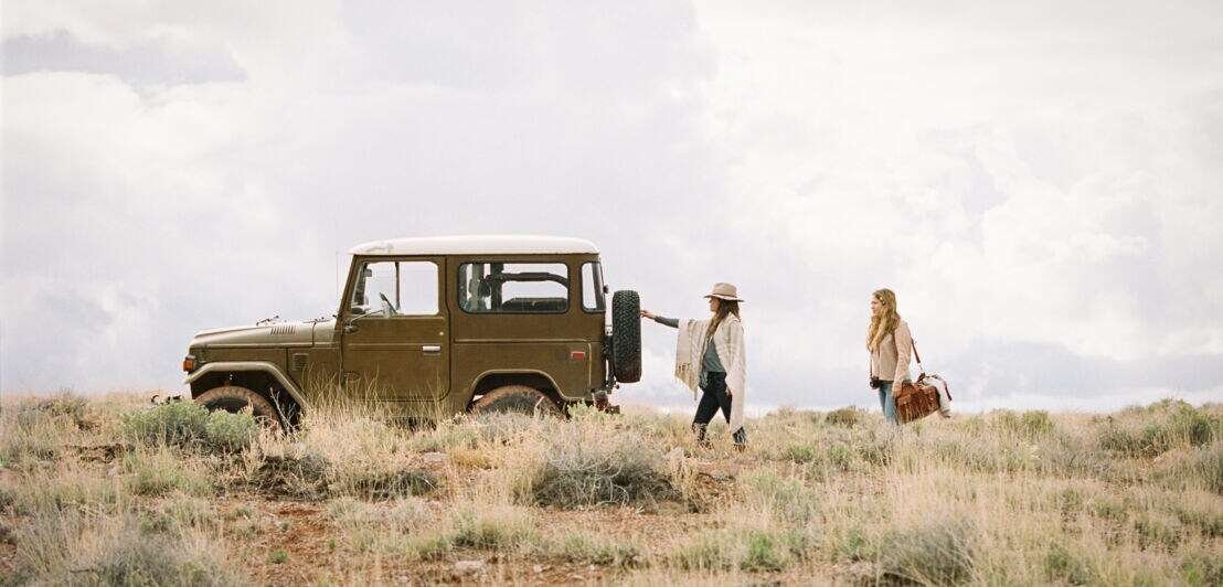 Zwei elegante Frauen mit Gepäck stehen während eines Urlaubstrips hinter einem Jeep in der Savanne